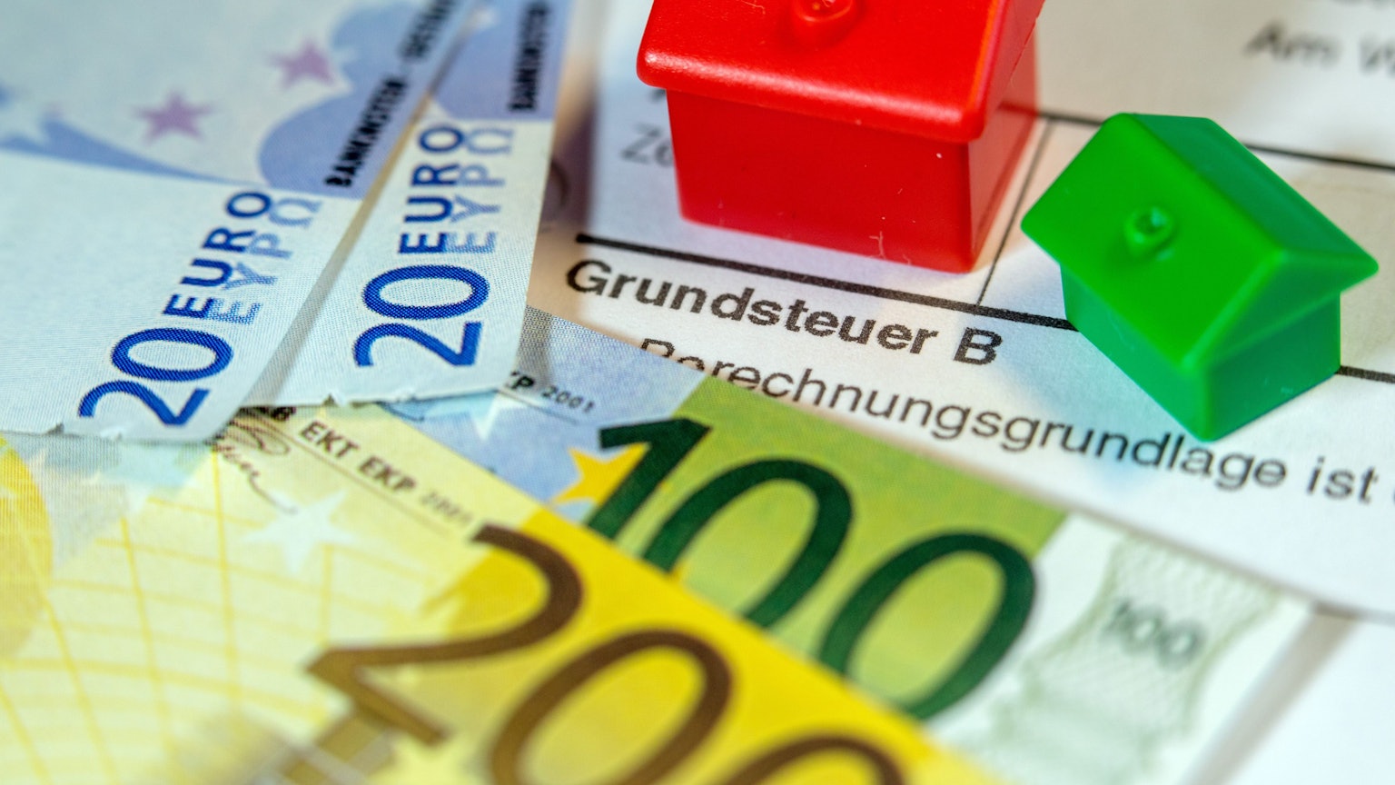 Ein Spielzeughaus und die Nachbildung von Euro-Geldscheinen liegen auf einem Abgabenbescheid für die Entrichtung der Grundsteuer.