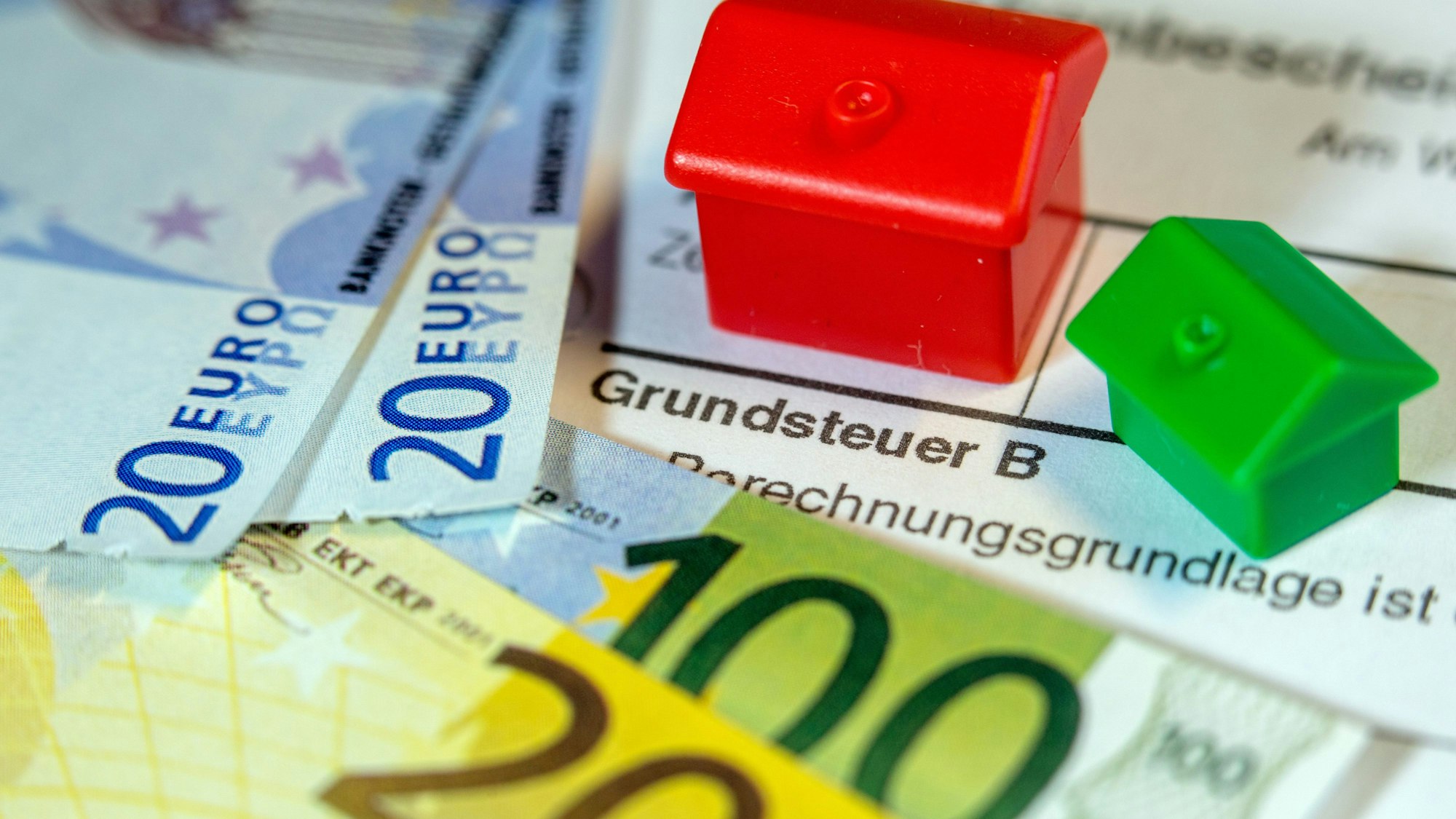 Zwei Monopoly-Häuser und die Nachbildung von Euro-Geldscheinen liegen auf einem Abgabenbescheid für die Entrichtung der Grundsteuer.