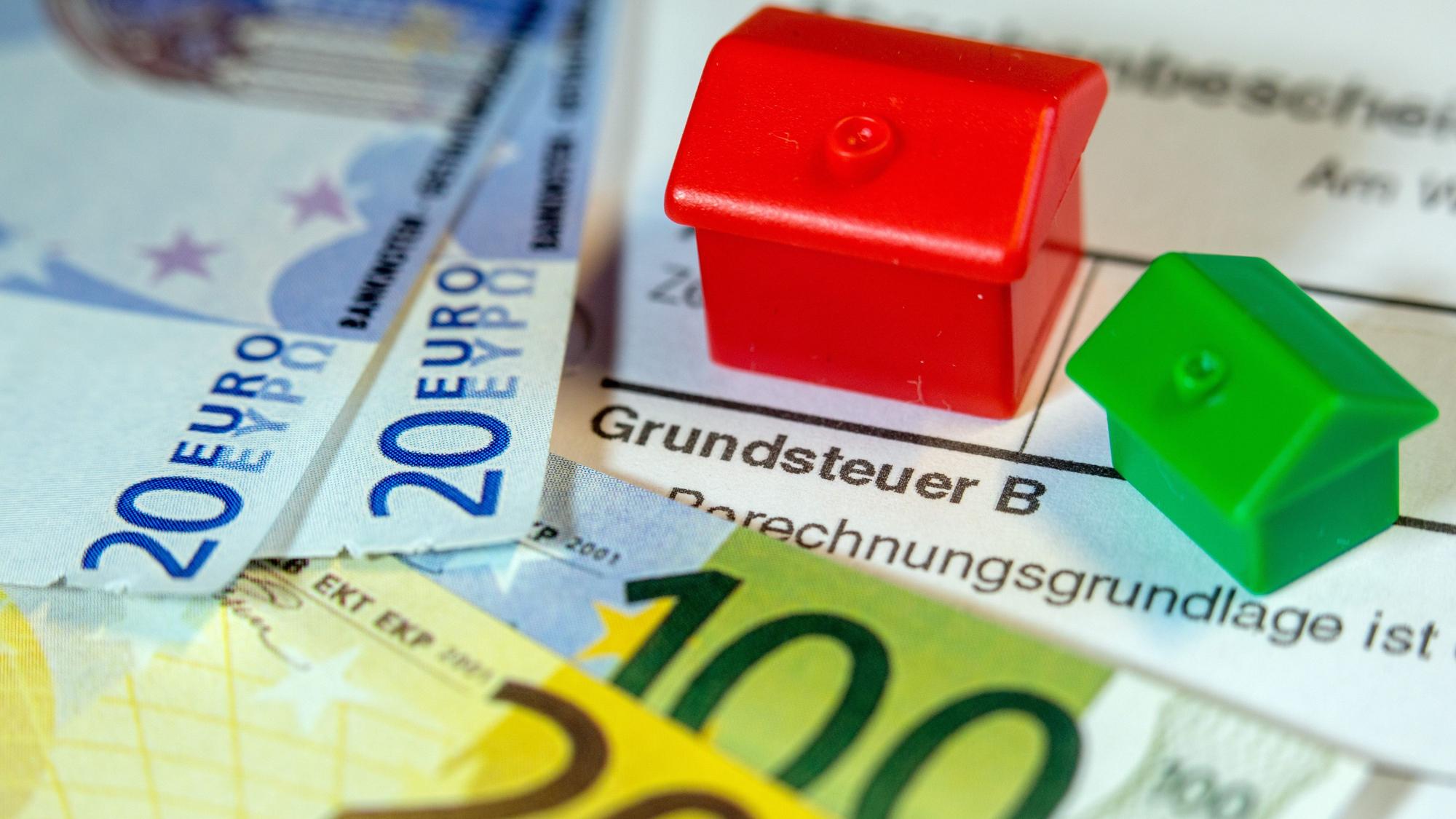 Ein Spielzeughaus und die Nachbildung von Euro-Geldscheinen liegen auf einem Abgabenbescheid für die Entrichtung der Grundsteuer.