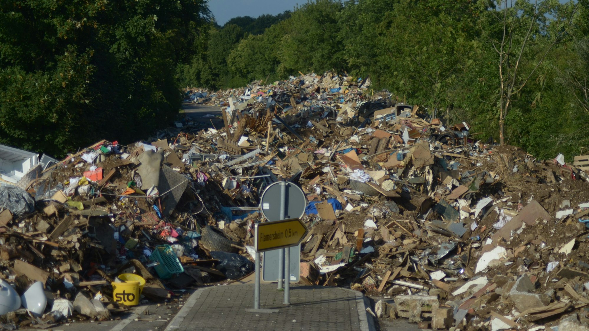 Große Mengen von Flutsperrmüll liegen auf einer von Bäumen gesäumten Straße.