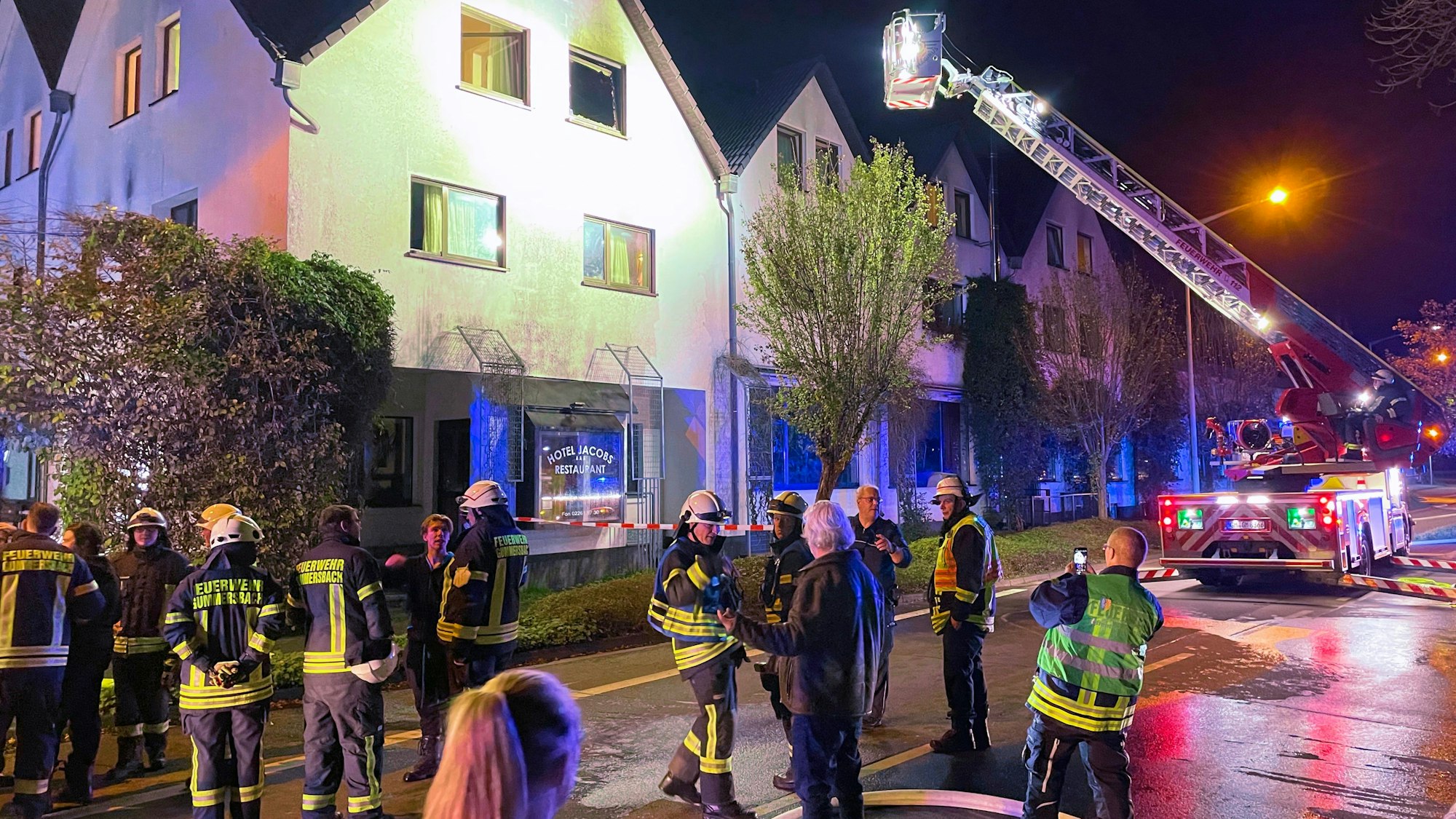 Zahlreiche Feuerwehrleute stehen vor dem Hotel Jacobs, im Hintergrund ist ein Feuerwehrwagen mit Drehleiter zu sehen.