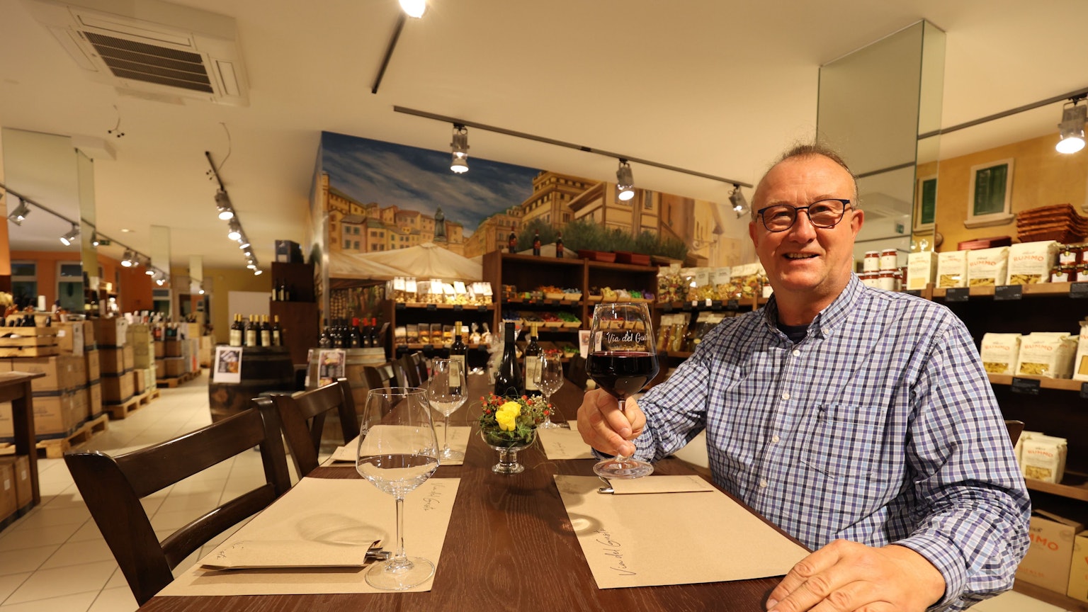 Betreiber des Ladens und Restaurants Via del Gusto in Rheinbach Jörg Meyer sitzt am gedeckten Tisch und hält ein gefülltes Rotweinglas in der Hand.