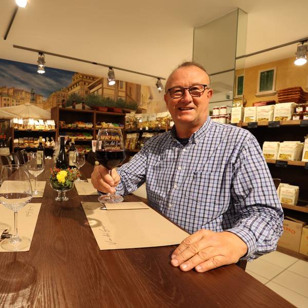 Betreiber des Ladens und Restaurants Via del Gusto in Rheinbach Jörg Meyer sitzt am gedeckten Tisch und hält ein gefülltes Rotweinglas in der Hand.