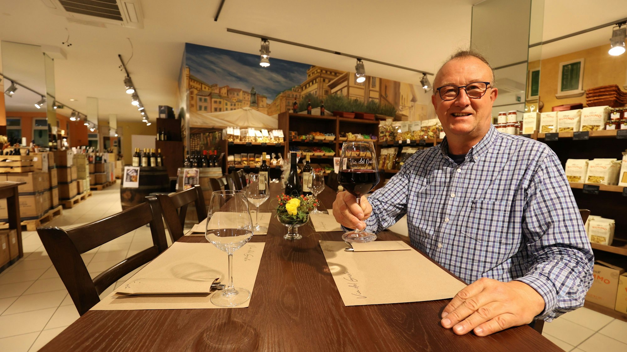 Betreiber des Ladens und Restaurants Via del Gusto in Rheinbach Jörg Meyer sitzt am gedeckten Tisch und hält ein gefülltes Rotweinglas in der Hand.