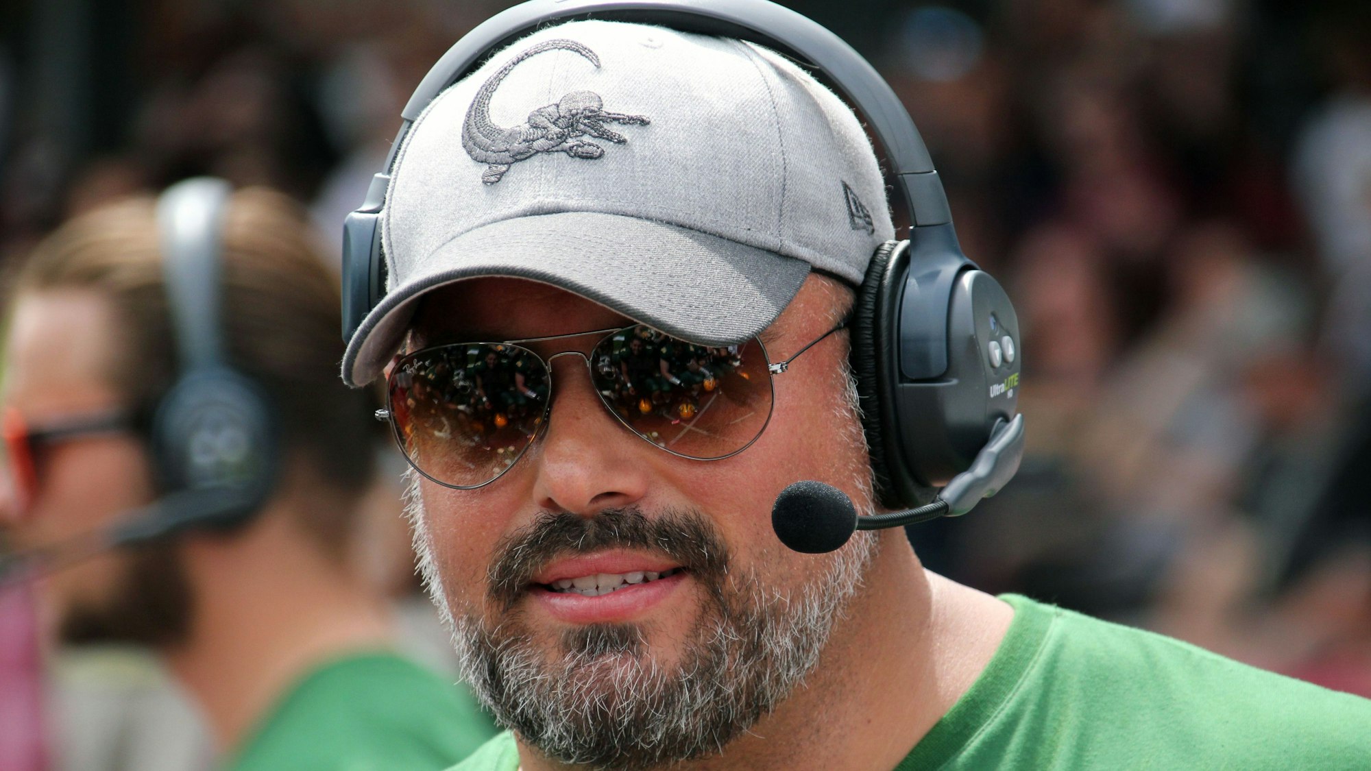 Ein Football-Trainer steht mit Kappe, Kopfhörern und Sonnenbrille am Spielfeldrand.