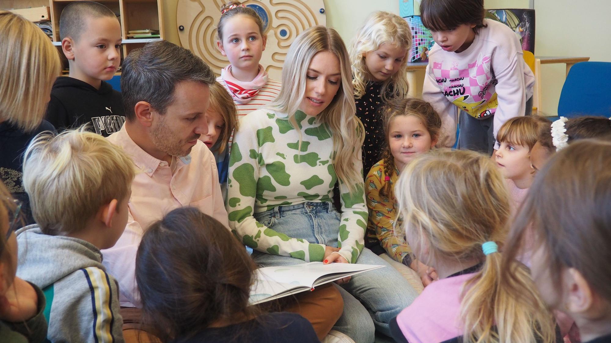 Marc Dumitru und Vanessa Meisinger sitzen in einem Kreis von Grundschulkindern und lesen aus Büchern vor.