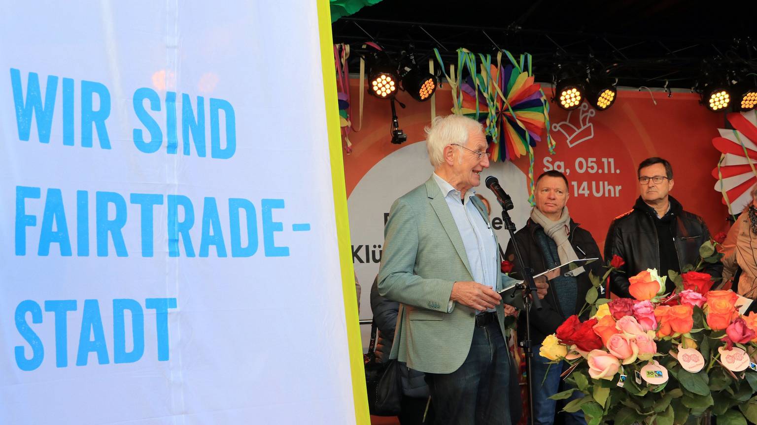 Auf einer Bühne stehen drei Männer hinter einer Flagge mit der Aufschrift „Wir sind Fair-Trade-Stadt“, unter anderem Ehrenbotschafter Manfred Holz am Mikrofon.
