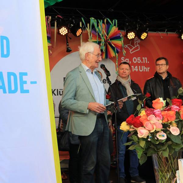 Auf einer Bühne stehen drei Männer hinter einer Flagge mit der Aufschrift „Wir sind Fair-Trade-Stadt“, unter anderem Ehrenbotschafter Manfred Holz am Mikrofon.