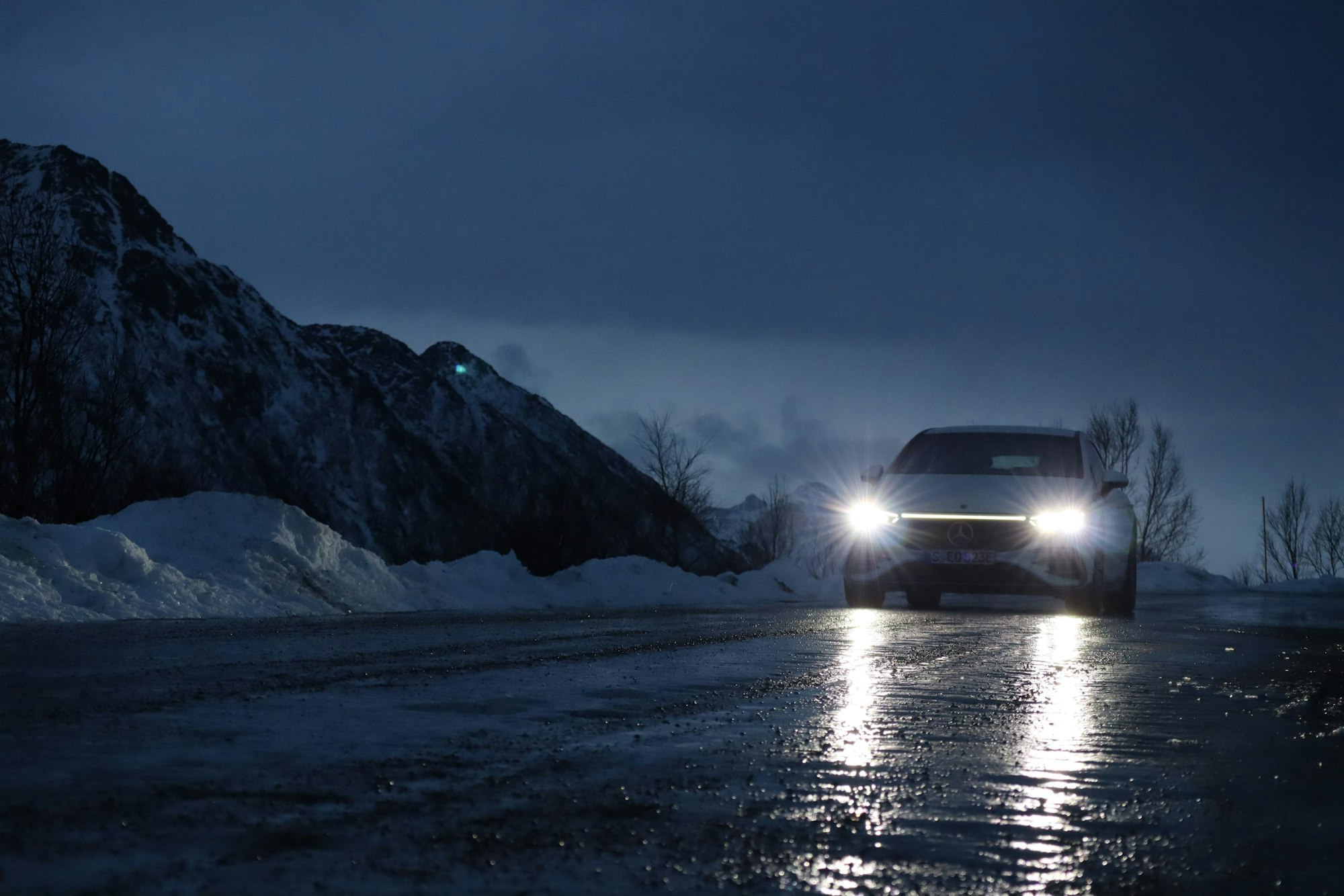 Ein E-Auto fährt im Halbdunkel über eine vereiste Straße im Winter, am Straßenrand liegt Schnee, im Hintergrund sind kahle Bäume zu sehen.