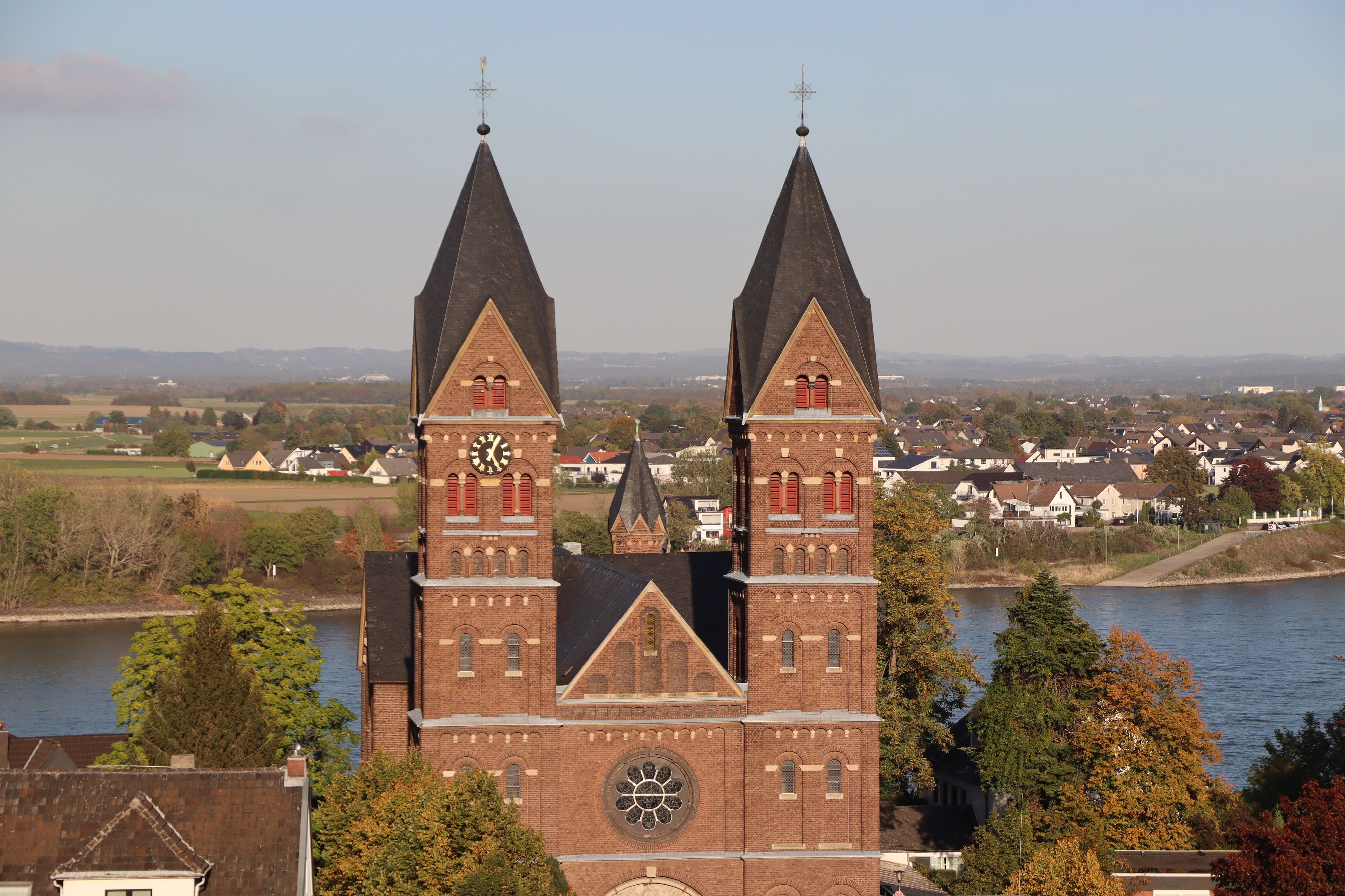 Die Kirchtürme der Pfarrkirche St. Germanus in Wesseling sind vor einem Gewässer zu sehen.