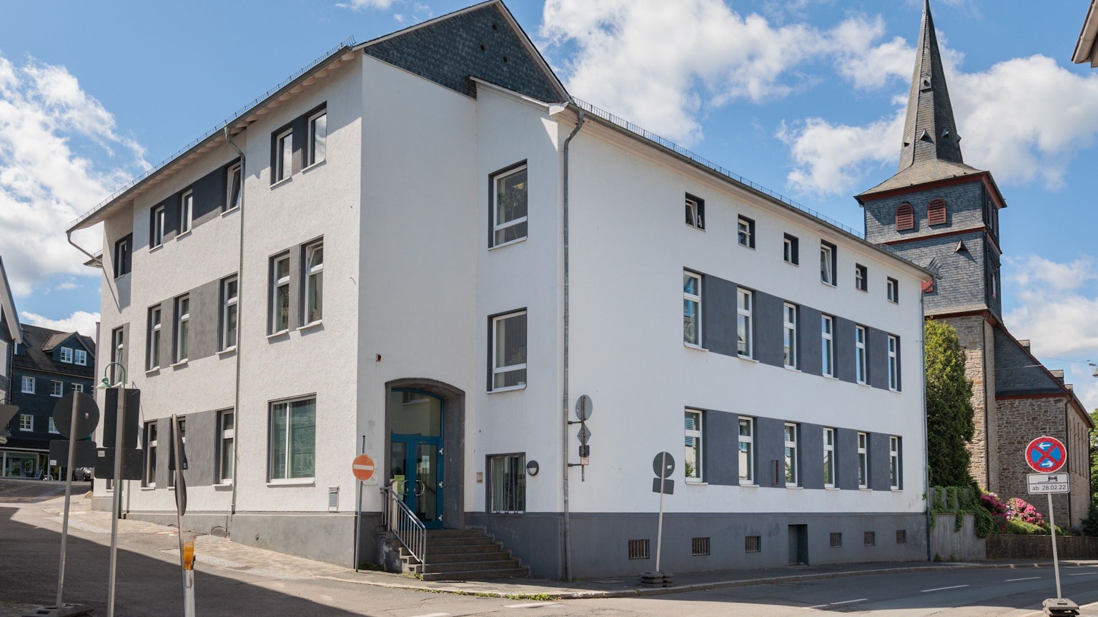 Das Bild zeigt ein weißes Gebäude, das Amtsgericht in Waldbröl. Im Hintergrund ist eine Kirche zu sehen.