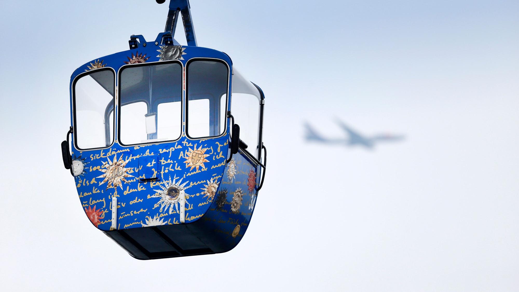 Die Gondel einer Seilbahn vor freiem Himmel, im Hintergrund ist undeutlich ein Flugzeug zu erkennen.