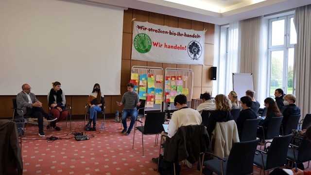 Mehrere Personen sitzen in einem Saal, im Hintergrund Plakate mit der Aufschrift „Wir handeln“.