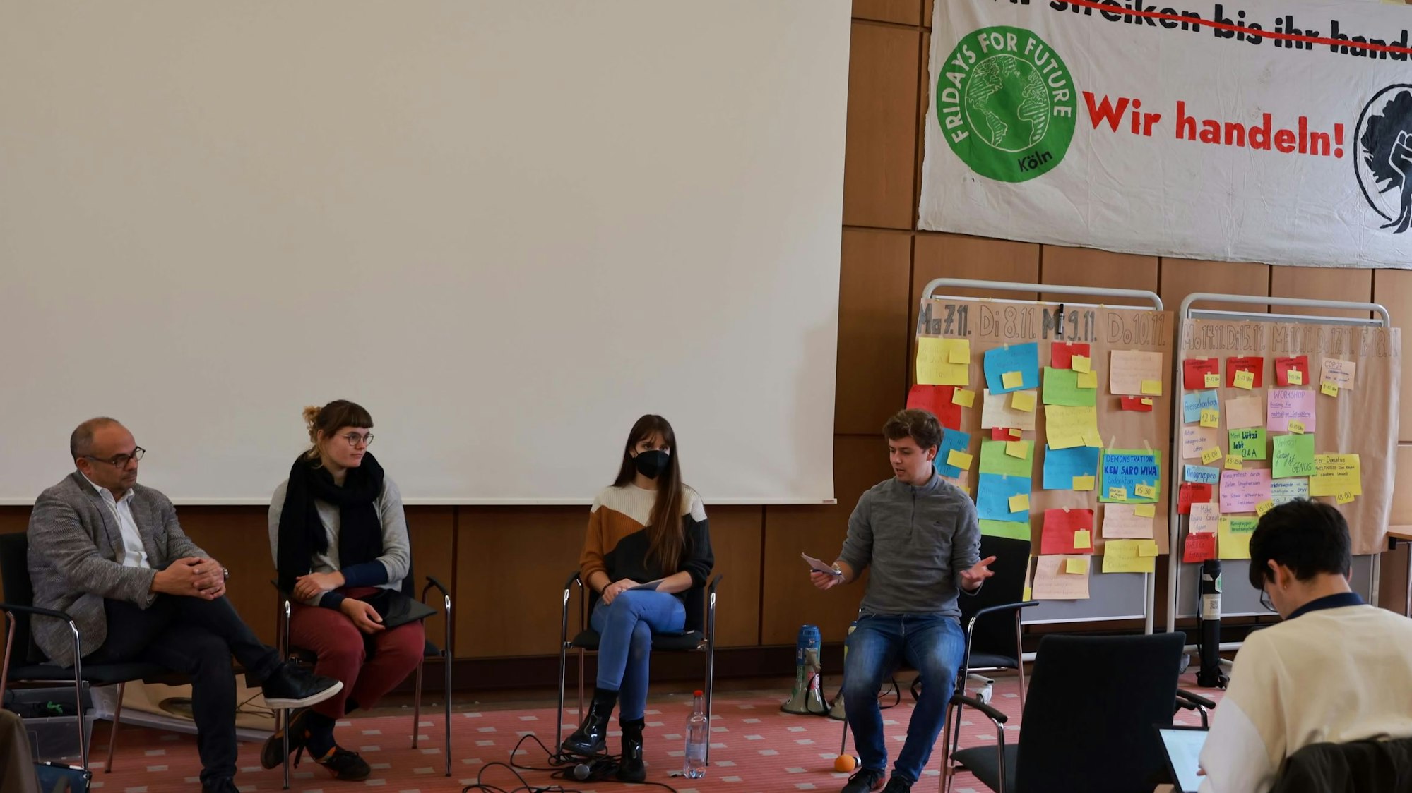 Mehrere Personen sitzen in einem Saal, im Hintergrund Plakate mit der Aufschrift „Wir handeln“.