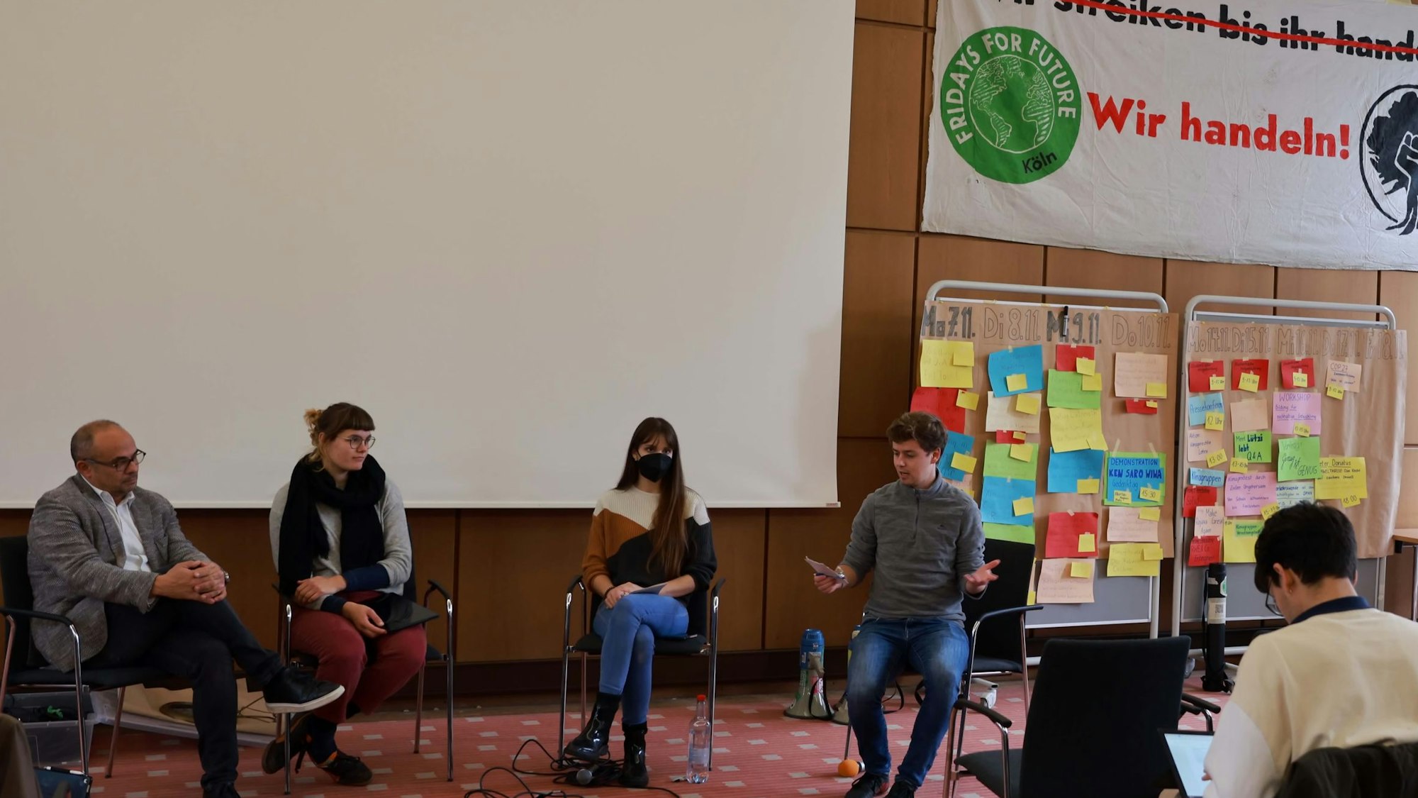 Mehrere Personen sitzen in einem Saal, im Hintergrund Plakate mit der Aufschrift „Wir handeln“.