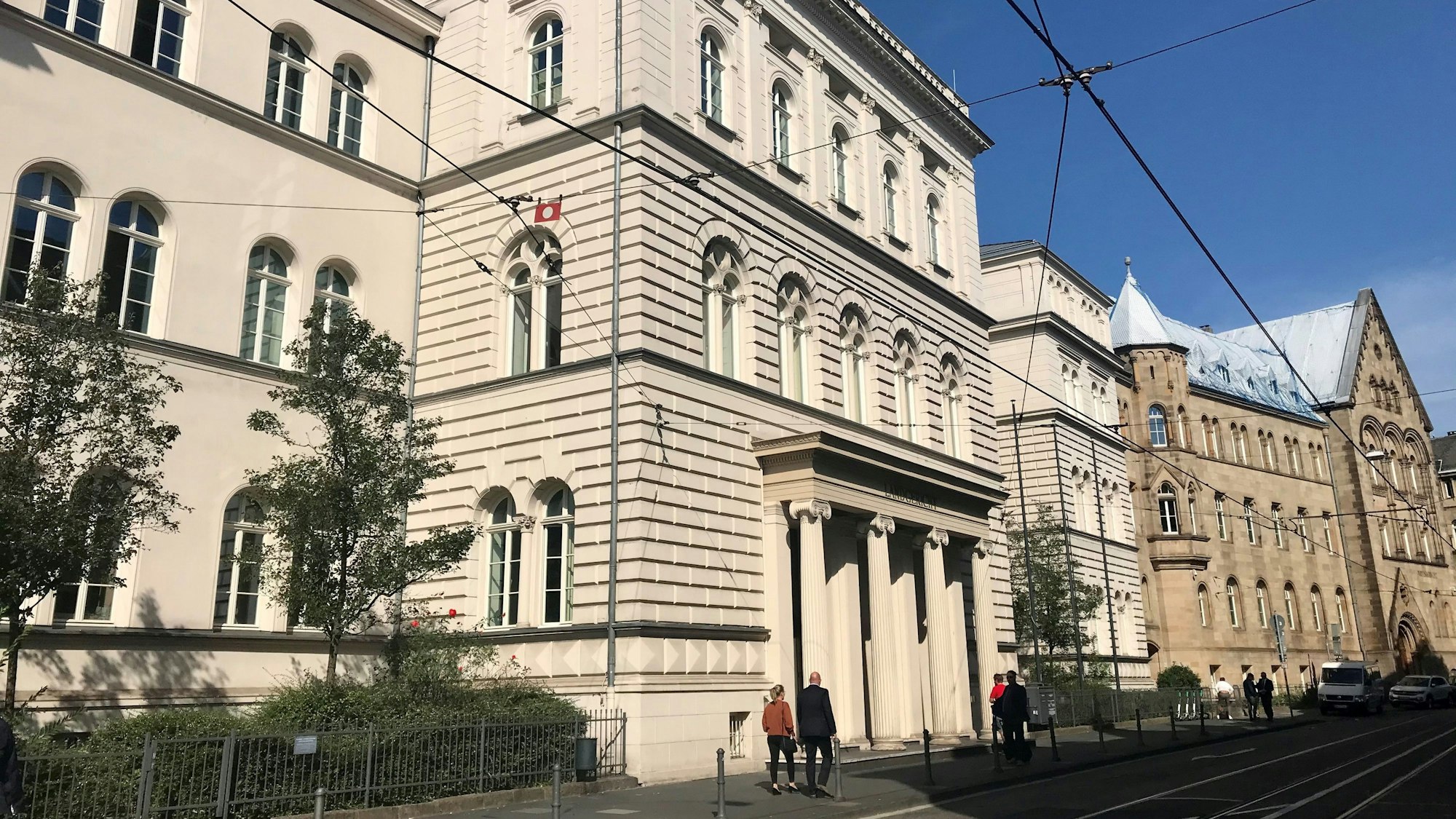 Ein großes helles Gebäude mit Säulen vor dem Eingang, Menschen gehen daran vorbei.