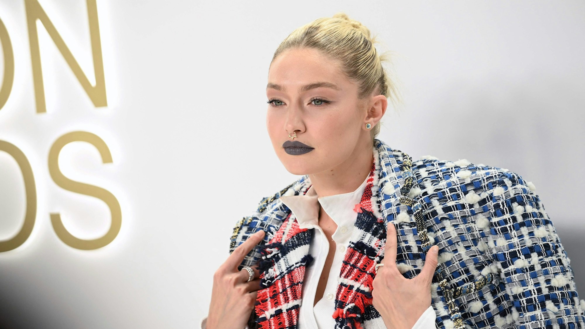Gigi hadid steht vor hellem Hintergrund und schaut an der Kamera vorbei. Sie hält mit ihren Händen ihren Mantel über den Schultern fest.