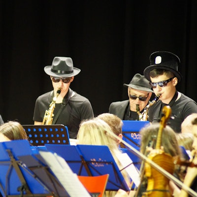 Männer mit Hüten und Sonnenbrillen spielen Blasinstrumente. Vor ihnen stehen Schulkinder mit ihren Notenständer.