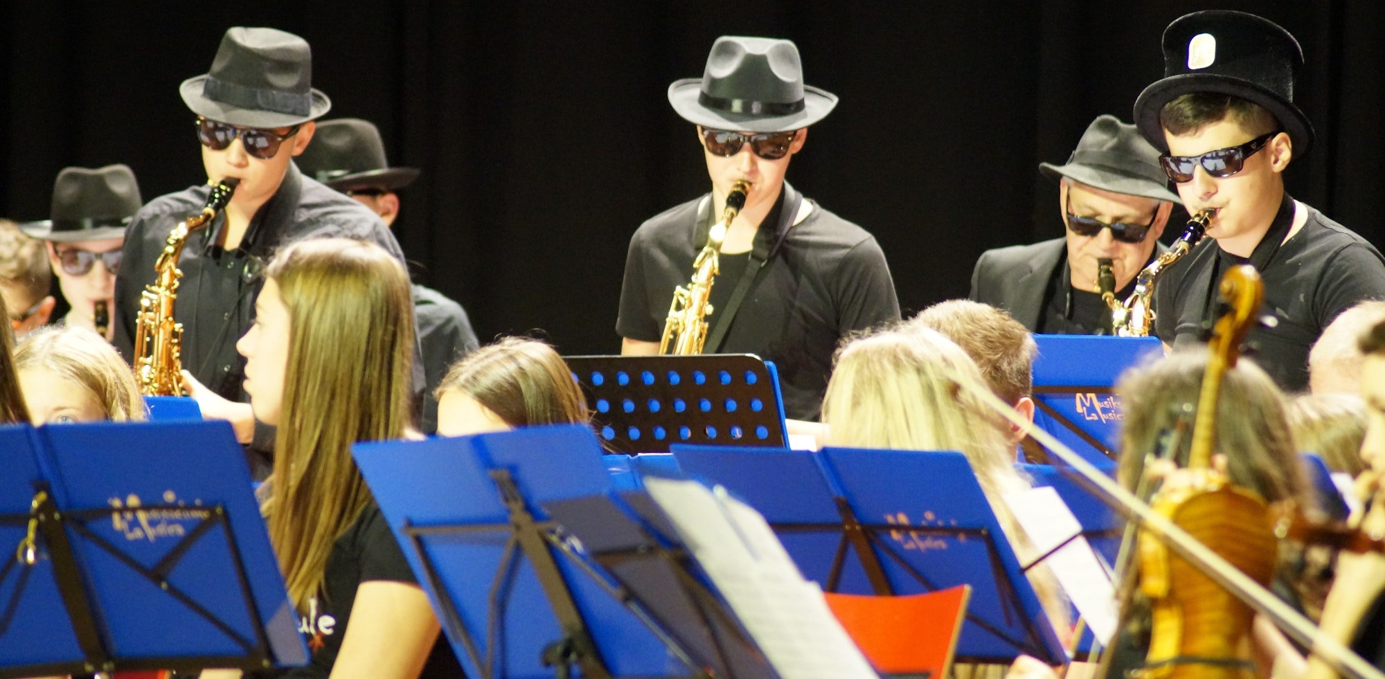 Männer mit Hüten und Sonnenbrillen spielen Blasinstrumente. Vor ihnen stehen Schulkinder mit ihren Notenständer.