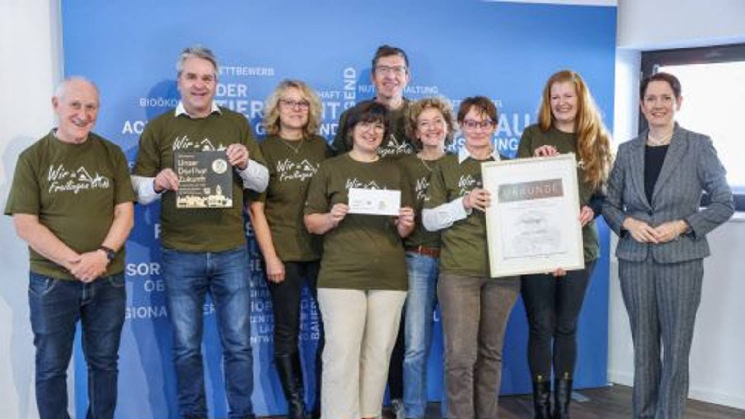 Eine Delegation von Preisträgern von „Unser Dorf hat Zukunft“ steht vor einer blauen Wand. Einige der Beteiligten halten Urkunden in die Kamera.