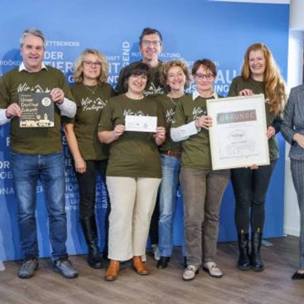 Eine Delegation von Preisträgern von „Unser Dorf hat Zukunft“ steht vor einer blauen Wand. Einige der Beteiligten halten Urkunden in die Kamera.