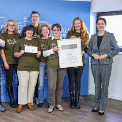 Eine Delegation von Preisträgern von „Unser Dorf hat Zukunft“ steht vor einer blauen Wand. Einige der Beteiligten halten Urkunden in die Kamera.