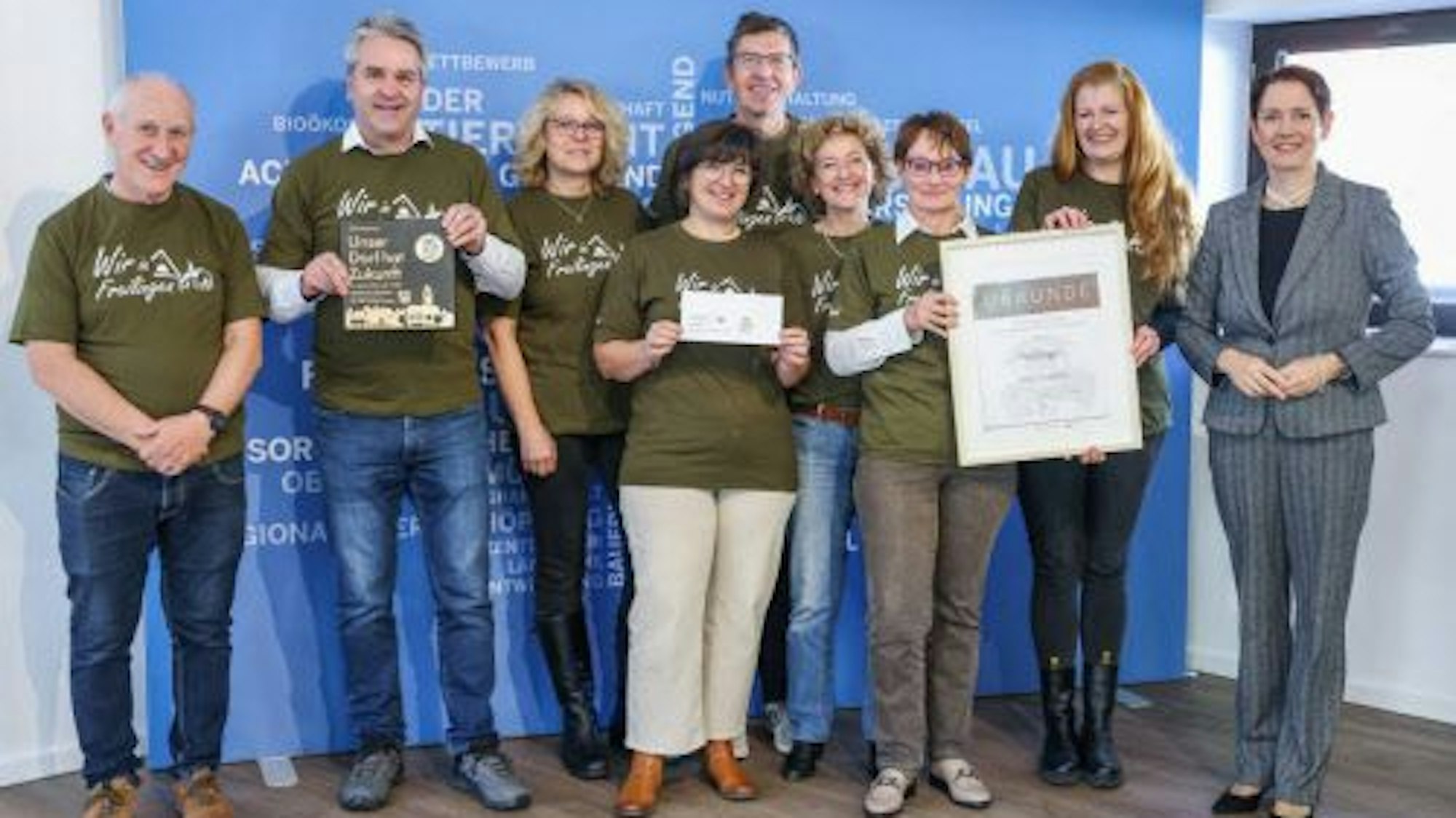 Eine Delegation von Preisträgern von „Unser Dorf hat Zukunft“ steht vor einer blauen Wand. Einige der Beteiligten halten Urkunden in die Kamera.