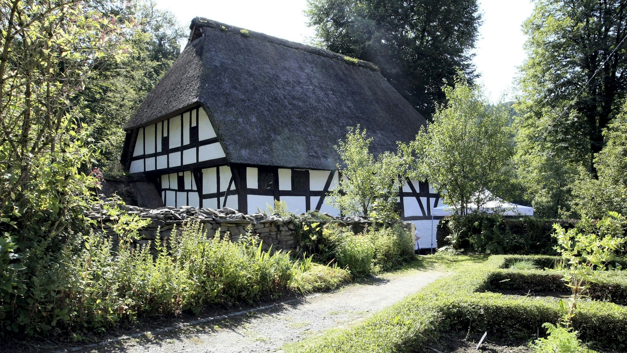Ein Fachwerkhaus mit einem Reetdach steht hinter einer Mauer. Im Vordergrund ist ein kleiner Weg und Hecken.
