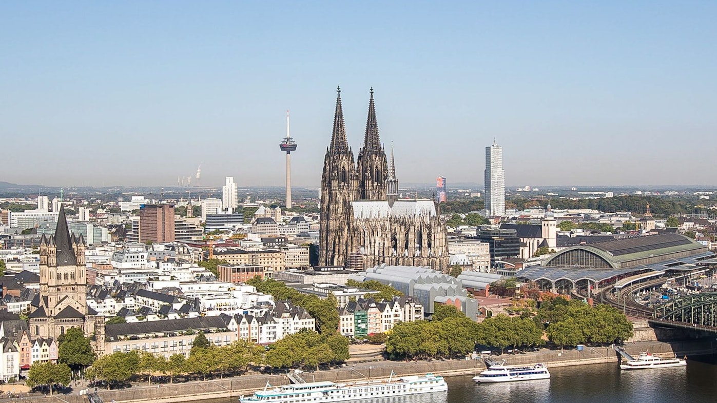 Köln mit Dom, Hohenzollernbrücke, Rhein und Hauptbahnhof bei strahlend blauem Himmel.