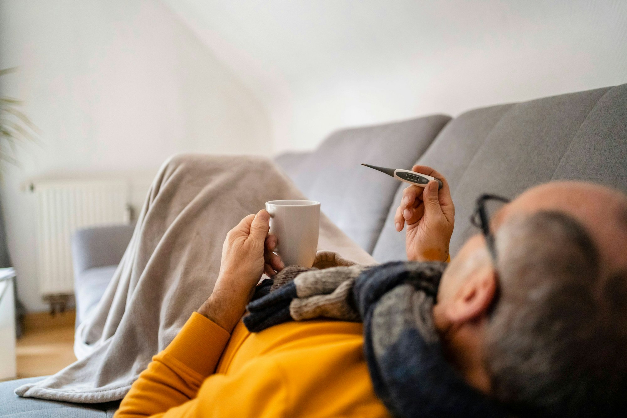 Ein Mann liegt krank auf einer Couch. Er hält eine Tasse und ein Fieber-Thermometer, seine Beine sind von einer Decke bedeckt.