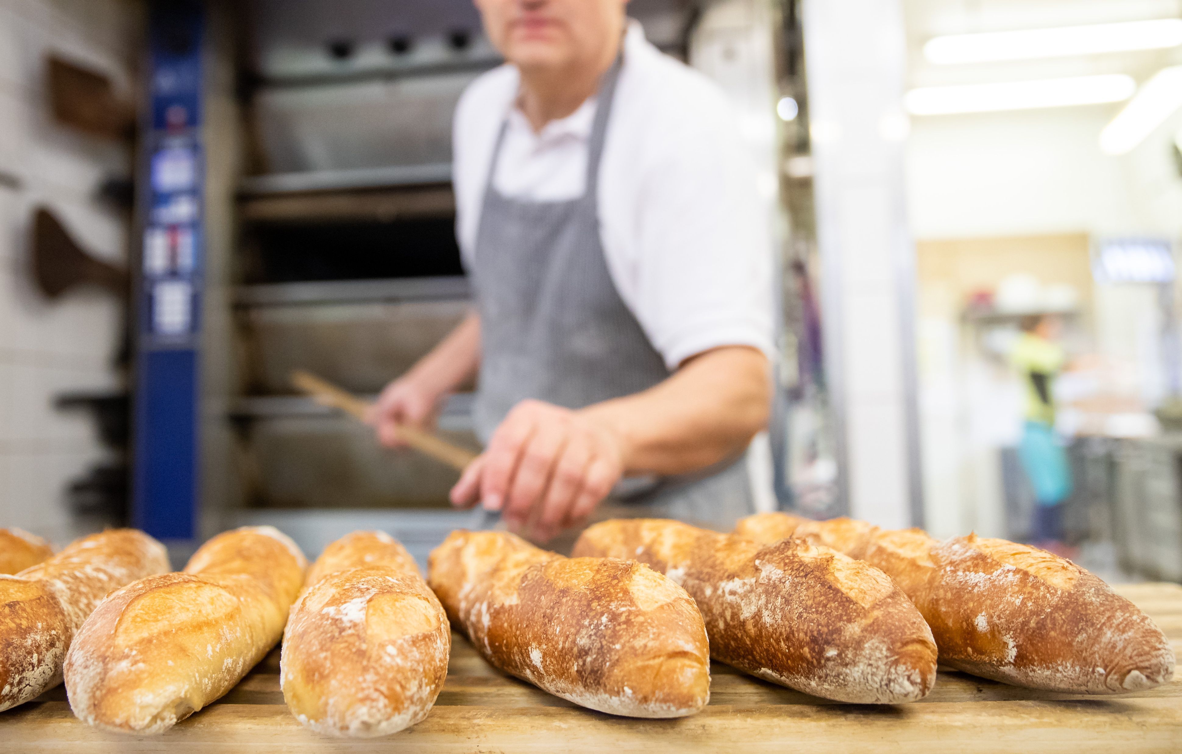 Mehrere Baguettes liegen auf einer Theke. Im Hintergrund steht ein Bäcker.