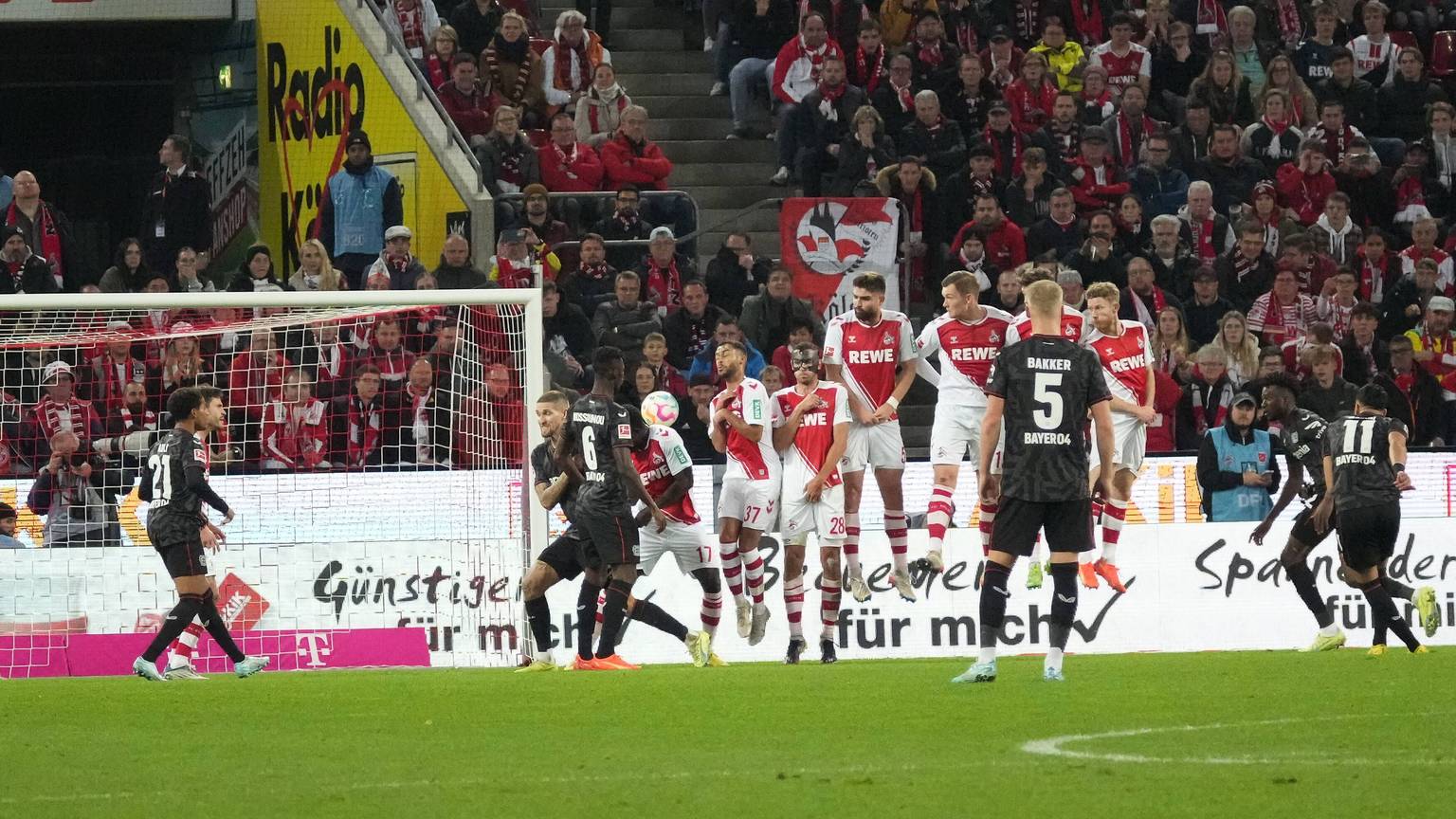 Auf dem Spielfeld ist links das Tor zu sehen, davor und daneben verschiedene Spieler beider Mannschaften. Der Ball befindet sich in der Mitte des Bildes.