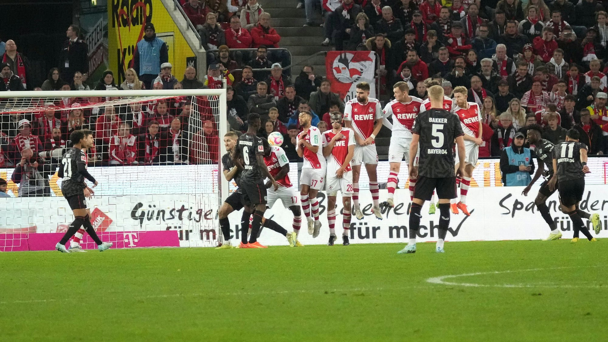Auf dem Spielfeld ist links das Tor zu sehen, davor und daneben verschiedene Spieler beider Mannschaften. Der Ball befindet sich in der Mitte des Bildes.