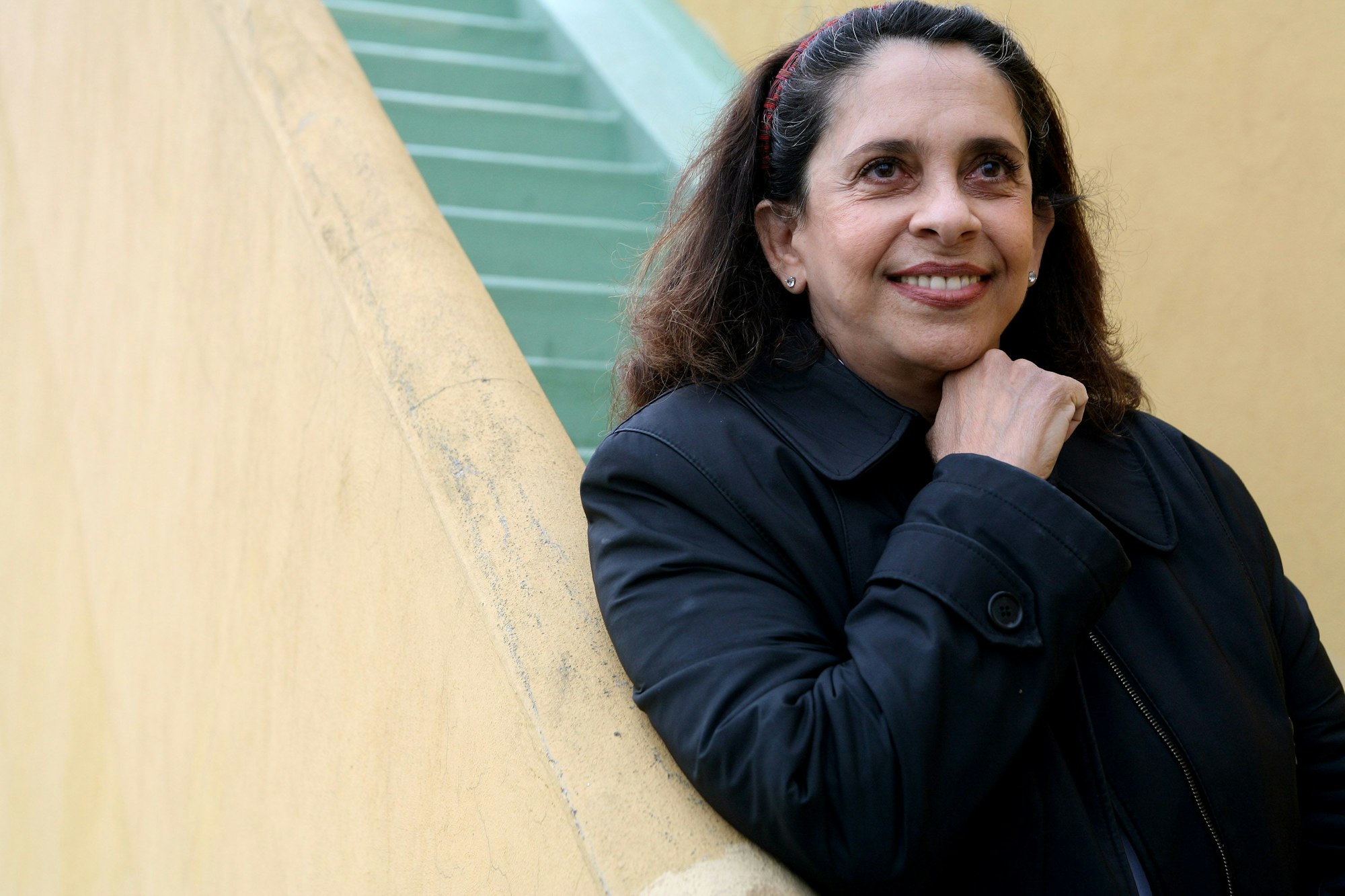 Gal Costa steht am Ende einer gelb-grünen Treppe. Sie hat einen dunkelblauen Mantel an und trägt die Haare offen. Sie lächelt und stützt die Hand am Geländer auf.