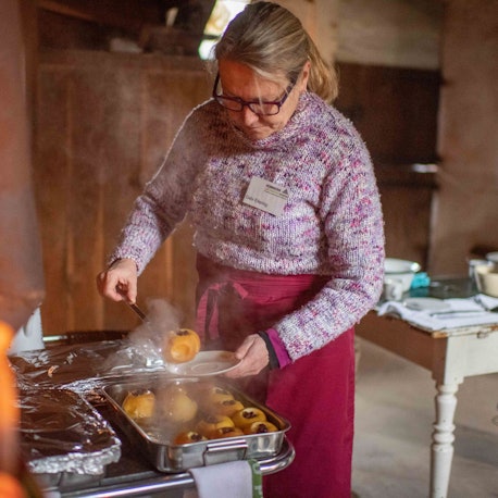 Eine Frau steht im Freilichtmuseum Kommern an einem Herd und hebt einen Bratapfel auf einen Teller.