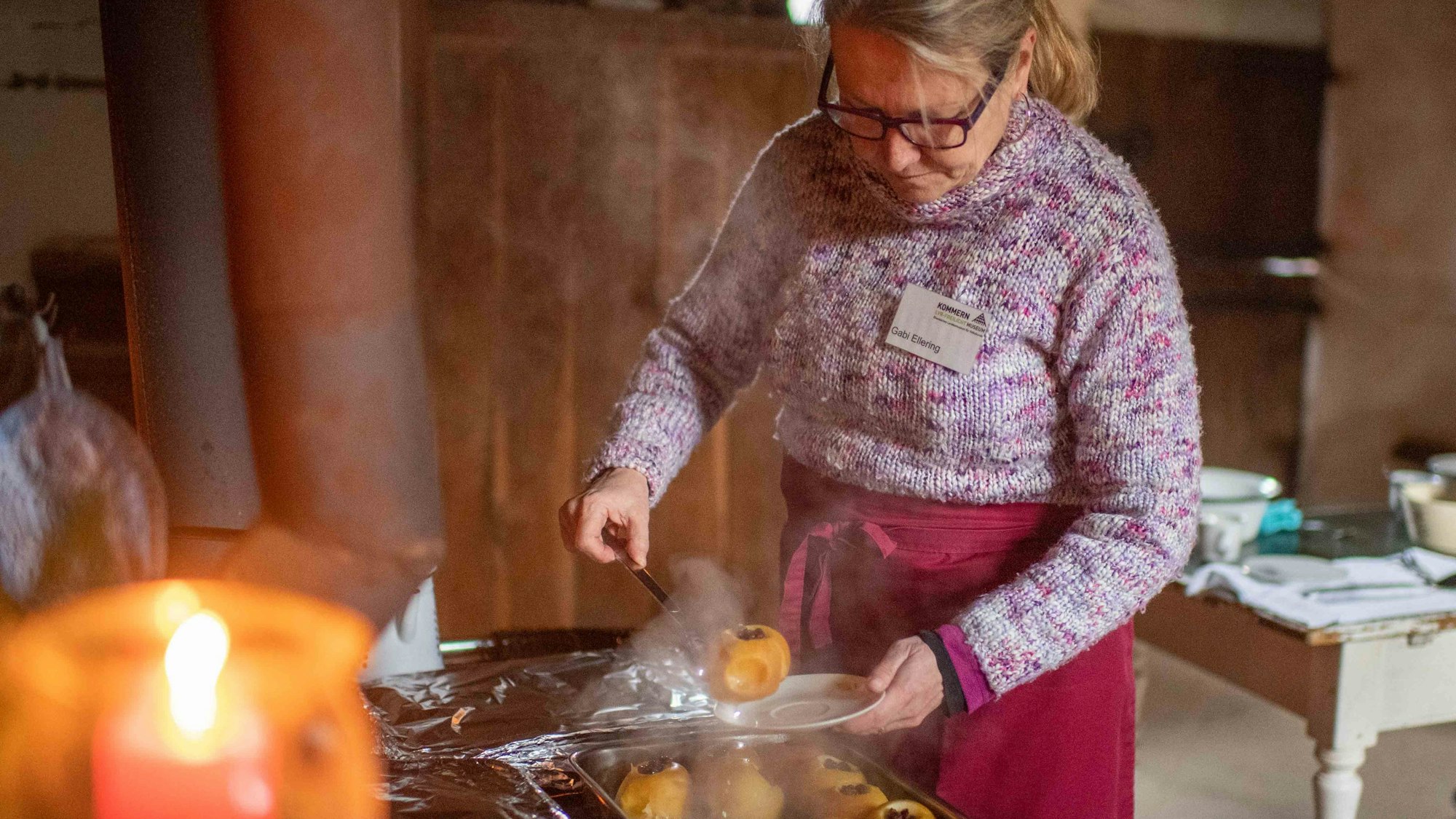 Eine Frau steht im Freilichtmuseum Kommern an einem Herd und hebt einen Bratapfel auf einen Teller.