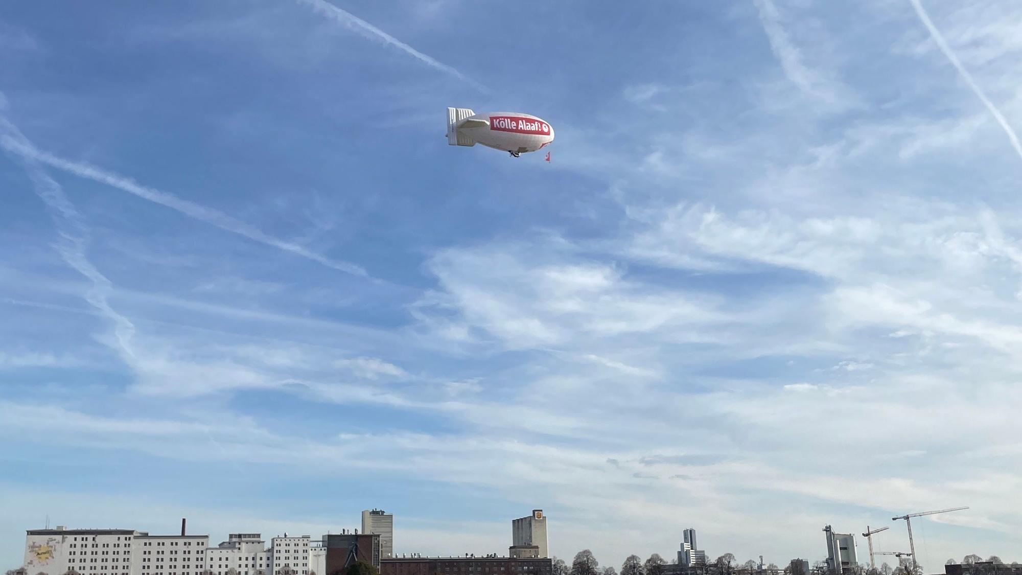 Bei strahlend blauem Himmel fliegt ein Zeppelin der Roten Funken mit der Aufschrift „Kölle Alaaf“ über den Rhein.