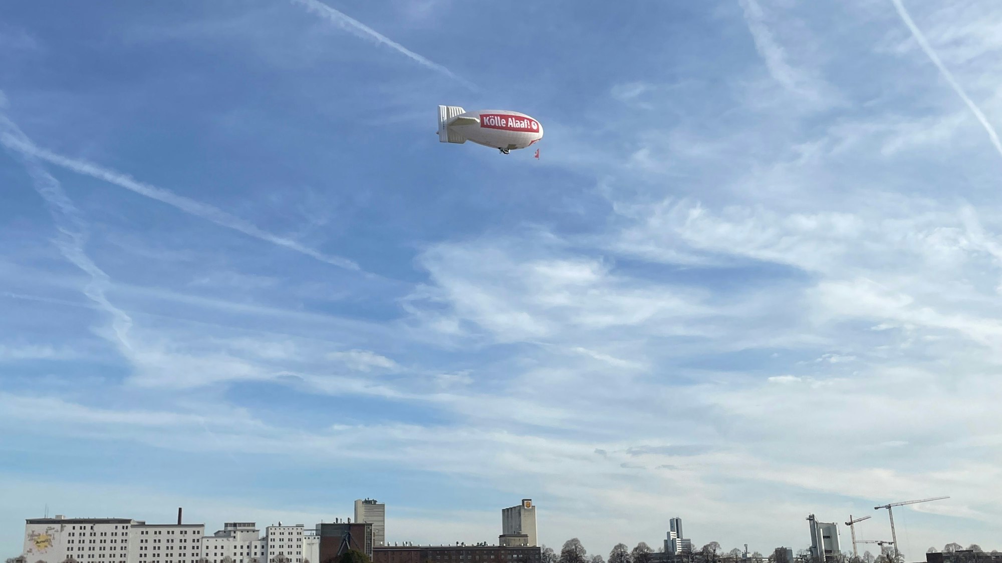 Bei strahlend blauem Himmel fliegt ein Zeppelin der Roten Funken mit der Aufschrift „Kölle Alaaf“ über den Rhein.
