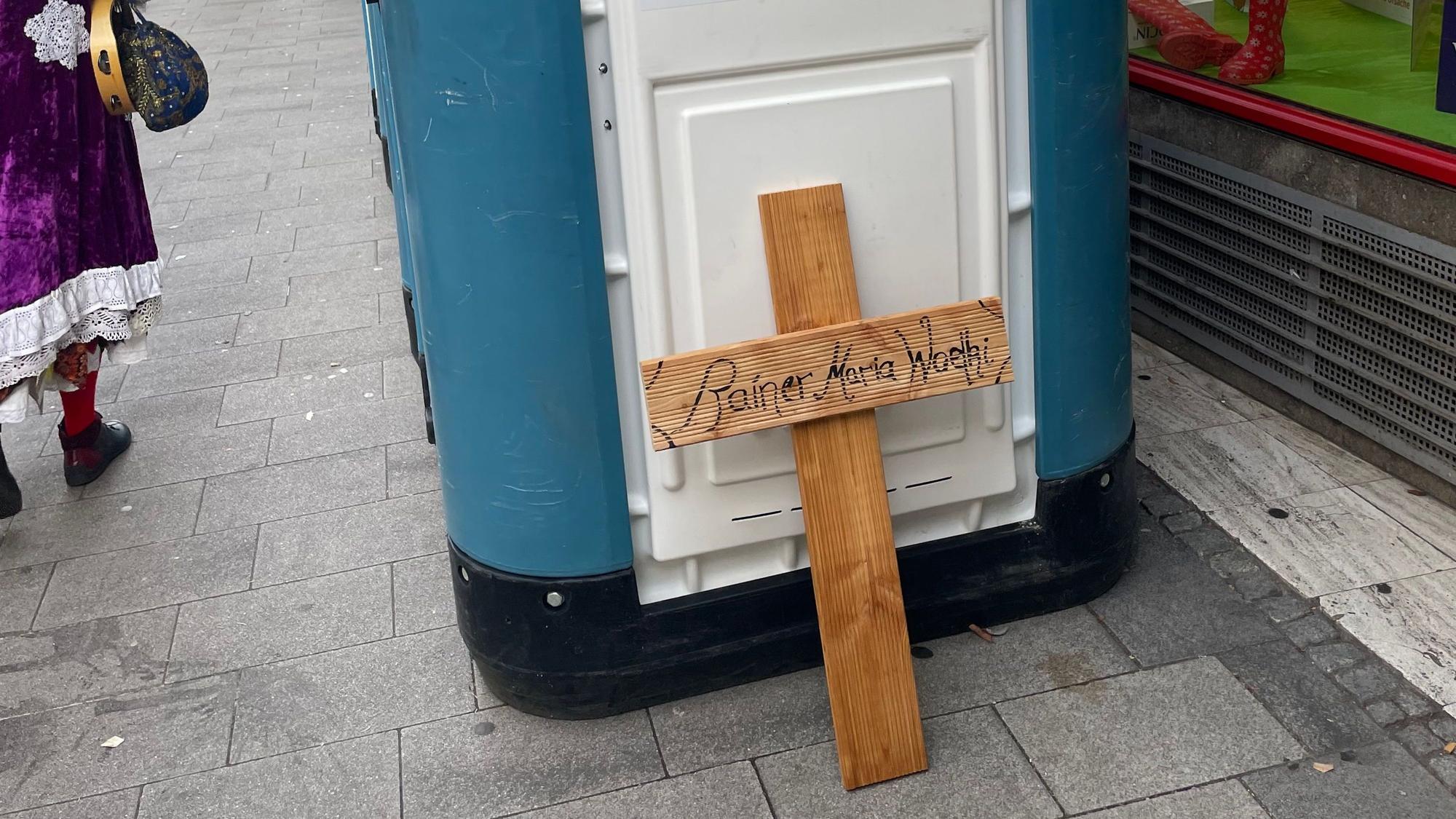Ein Holzkreuz mit der Aufschrift „Rainer Maria Woelki“ wurde an ein Dixiklo gelehnt.