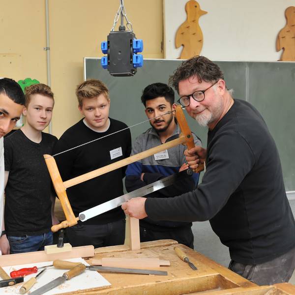 Ausbilder Arnulf Rückamp steht mit seinen Lehrlingen um einen Tisch mit Werkzeug und sägt Holz.