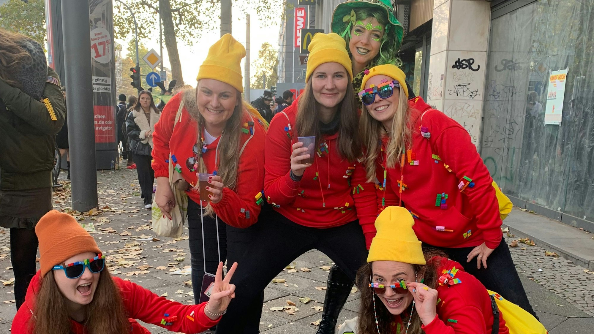 Fünf lachende Menschen, die schwarze Hosen, rote Pullover und gelbe Mützen tragen. Auf den Pullovern kleben Legosteine. Im Hintergrund eine weitere Person in grün.