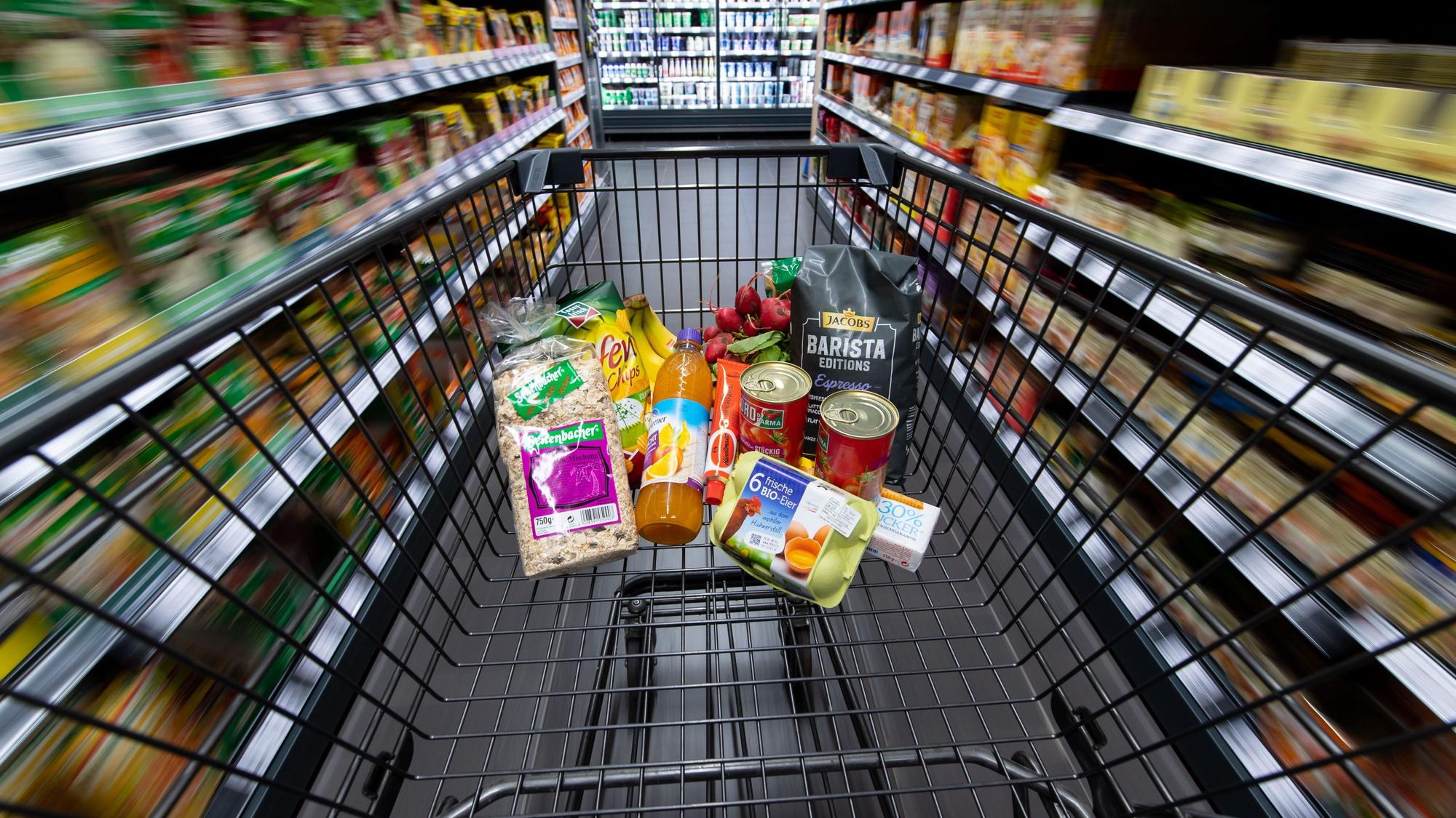Ein Einkaufswagen, gefüllt mit Konserven, Eiern und einigen anderen Lebensmitteln, wird durch einen Gang im Supermarkt geschoben.