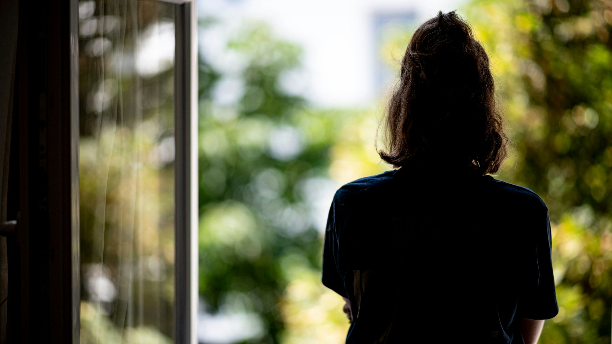 Man sieht eine Frau an einem geöffneten Fenster stehen. Sie dreht der Kamera den Rücken zu. Das Fenster zeigt ein helles grünes Äußeres, die Frau wirkt dagegen wie ein dunkler Kontrast.