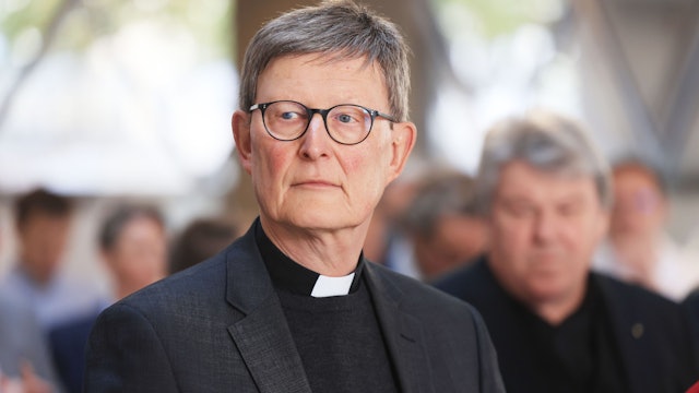 Kardinal Woelki blickt in die Kamera. Er trägt einen grauen Anzug und eine Brille, im Hintergrund sind Menschen zu sehen.