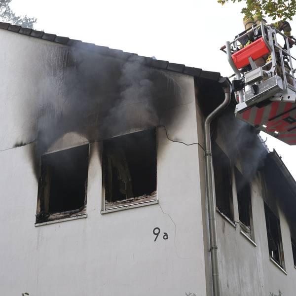 Ein Feuerwehrmann steht in einem Korb auf der Feuerwehrleiter. Die Hausfassade ist an mehreren Fenstern von dem Feuer zerstört.