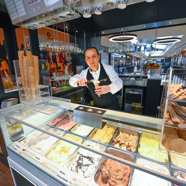 Ein Mann steht hinter der Theke einer Eisdiele und drücke eine Kugel Eis in eine Waffel. In der Auslage liegen verschiedene Geschmacksrichtungen.