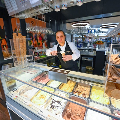 Ein Mann steht hinter der Theke einer Eisdiele und drücke eine Kugel Eis in eine Waffel. In der Auslage liegen verschiedene Geschmacksrichtungen.