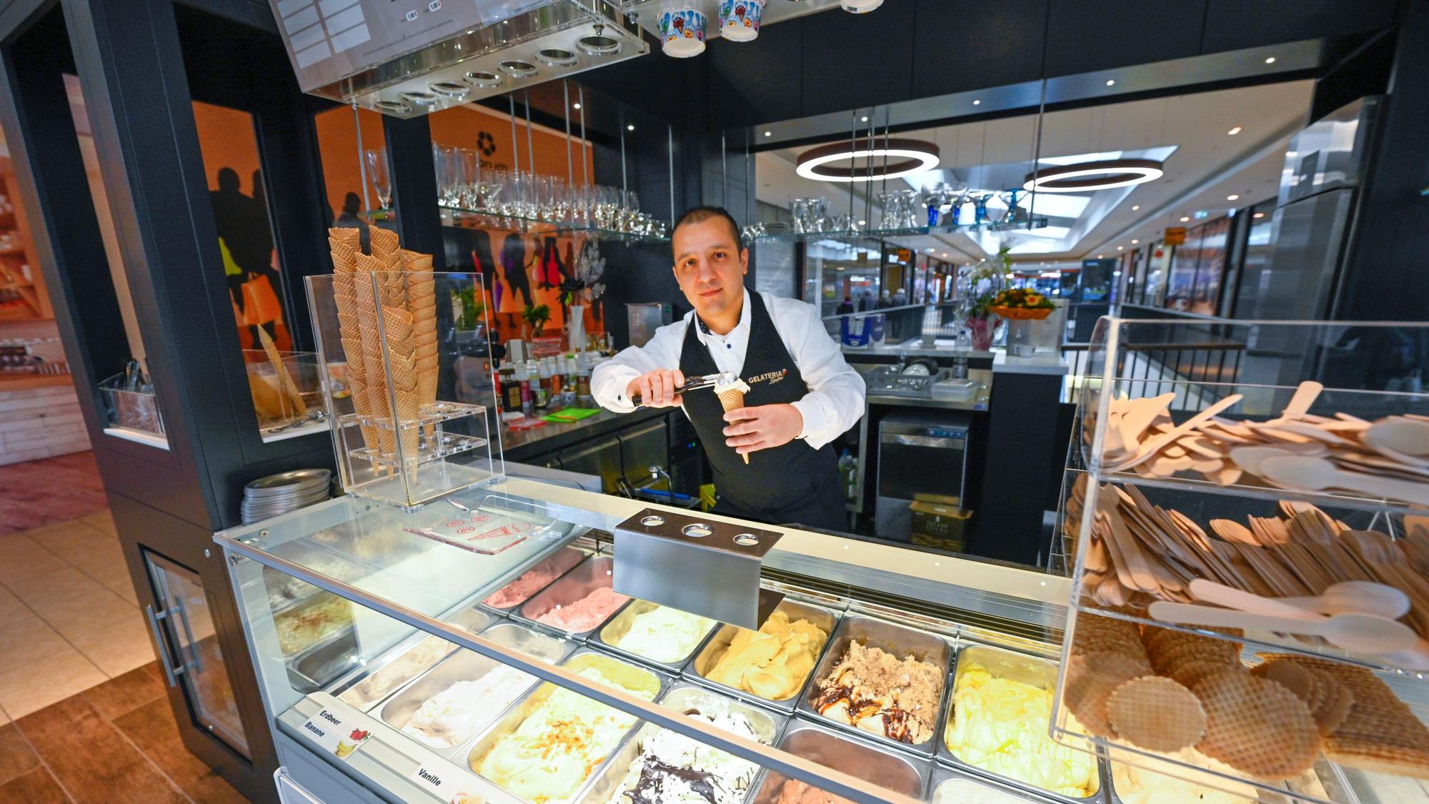Ein Mann steht hinter der Theke einer Eisdiele und drücke eine Kugel Eis in eine Waffel. In der Auslage liegen verschiedene Geschmacksrichtungen.