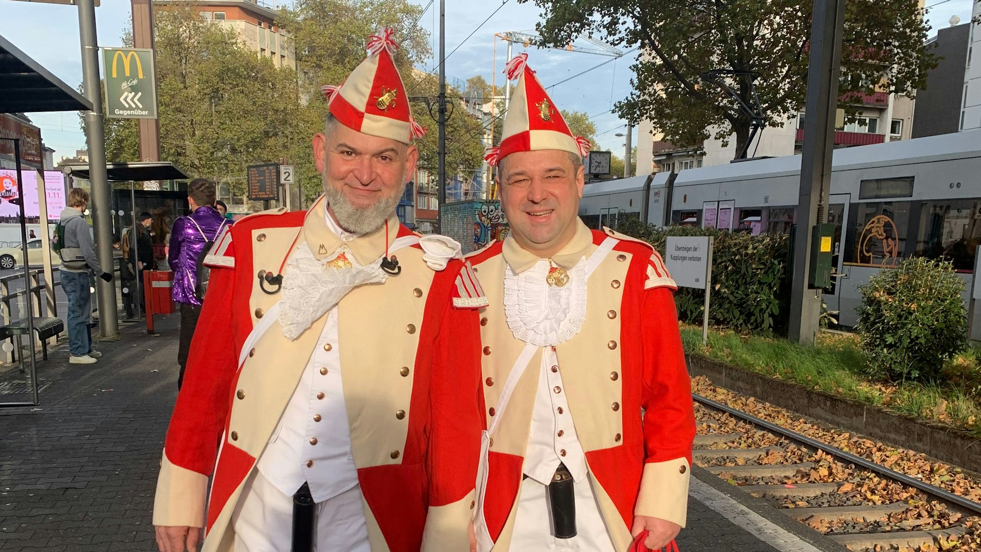 Zwei Menschen im Kostüm der roten Funken warten gut gelaunt am Bahnsteig.