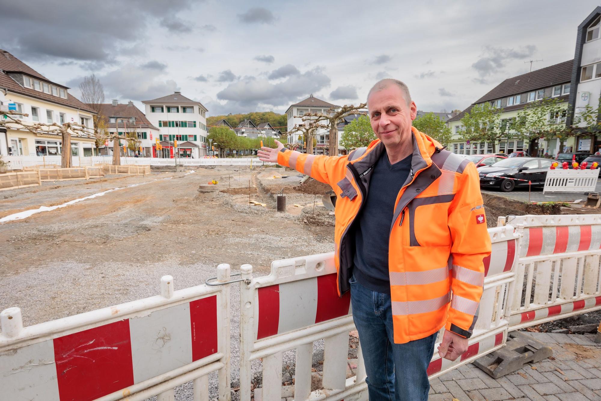 Andreas Kiel, Leiter der Abteilung Tiefbau bei der Gemeinde Engelskirchen, steht in einer signalorangefarbenen Jacke vor einer Warnbake am Bahnhofsplatz in Engelskirchen. Er weist im Bereich des Bahnhofs auf die Baustelle, die sich bis zur Märkischen Straße erstreckt. Die Hälfte des Platzes ist momentan Baustelle. Im Hintergrund sind die Platanen zu sehen, die stehen bleiben sollen.